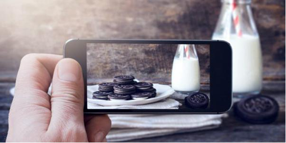 man using his mobile phone to take a photo of a stack of oreos on a plate with a glass bottle of milk at the side