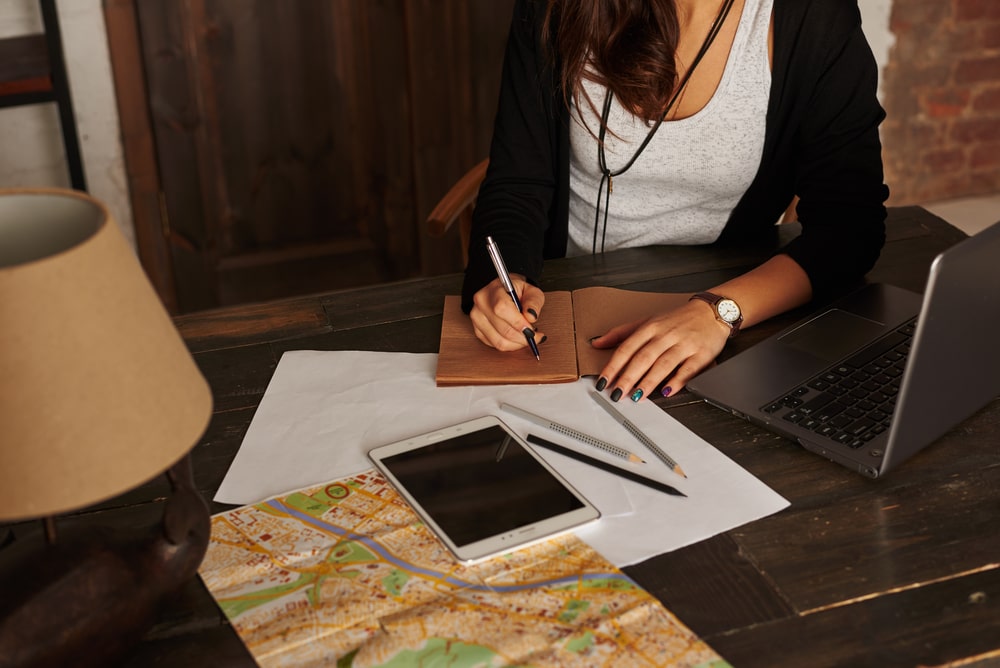 Woman planning new trip. Girl write tourist notes at home. Tablet, laptop, city map on the table. Girl learn the route in the new city. Loft interior with big wooden doors.