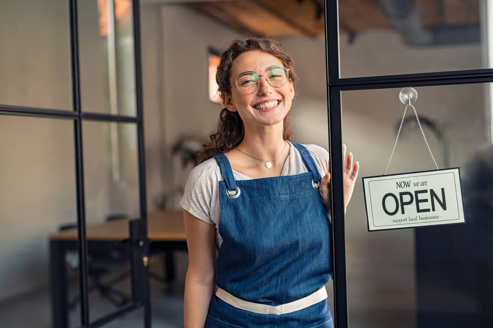 smiling local female shop owner standing at the entrance beside “OPEN” sign